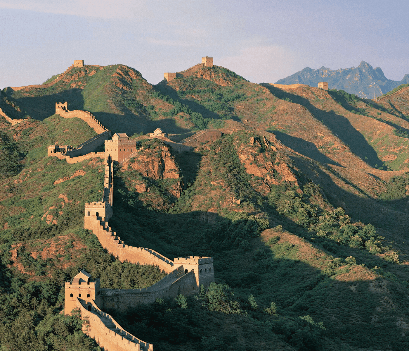 A sweeping aerial view of the Great Wall of China snaking across rolling, forested mountains under a clear blue sky, with sunlit stone watchtowers and fortified sections casting long shadows across the lush green and earth-toned hillsides, evoking the grandeur and historical scale of this iconic landmark.
